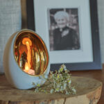 Ein LED-Teelicht (wiederaufladbar, mit Fernbedienung, wasserdicht, bis zu 400 Std. Brenndauer) leuchtet in einem weißen und goldenen Halter auf Holz, umgeben von weißen Blumen; im Hintergrund ist ein unscharfes Porträt zu sehen.