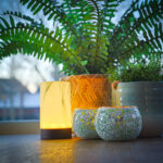 Zwölf warmweiße wiederaufladbare LED-Teelichter (bis zu 400h Brenndauer, wasserdicht, mit Fernbedienung) sind auf einer Fensterbank angeordnet, im Hintergrund sind Bäume und Gebäude zu sehen.