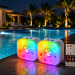 Twee vierkante, veelkleurige LED zwembadlampen met afstandsbediening en USB-kabel zijn 's nachts aan de rand van een verlicht zwembad geplaatst, met palmbomen en een modern huis op de achtergrond.