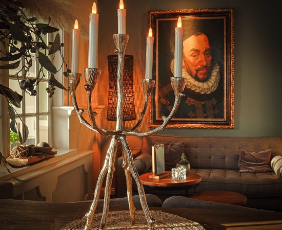 Een zilveren kandelaar met zes LED dinerkaarsen oplaadbaar 97mm 130 uur warm wit (set van 6, met afstandsbediening) staat op een geweven mat in een gezellige kamer en werpt warm licht bij een bank en een groot ingelijst portret.