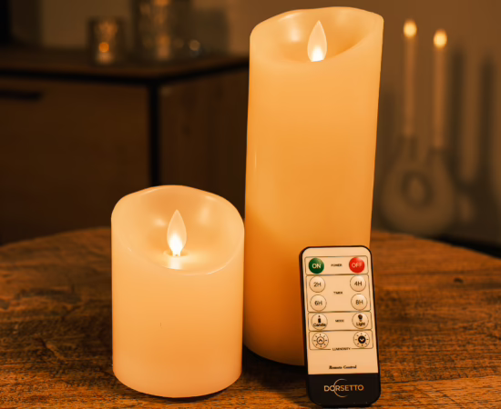 Een set van twee oplaadbare led kaarsen van Dorsetto, een hoge en een korte, staan op een houten tafel met een afstandsbediening met Dorset-label ervoor en een zacht verlichte, onscherpe achtergrond.