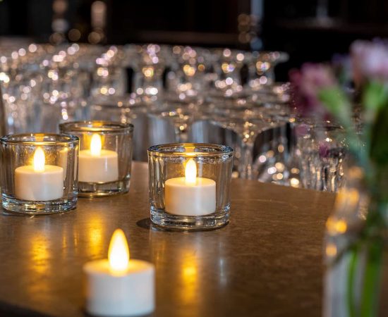 Twaalf Led waxinelichtjes waterdicht oplaadbaar brandtijd 175 uur warm wit met afstandsbediening staan in heldere houders op een aanrecht, met onscherp glaswerk en bloemen op de achtergrond, waardoor een warme, uitnodigende sfeer wordt gecreëerd.