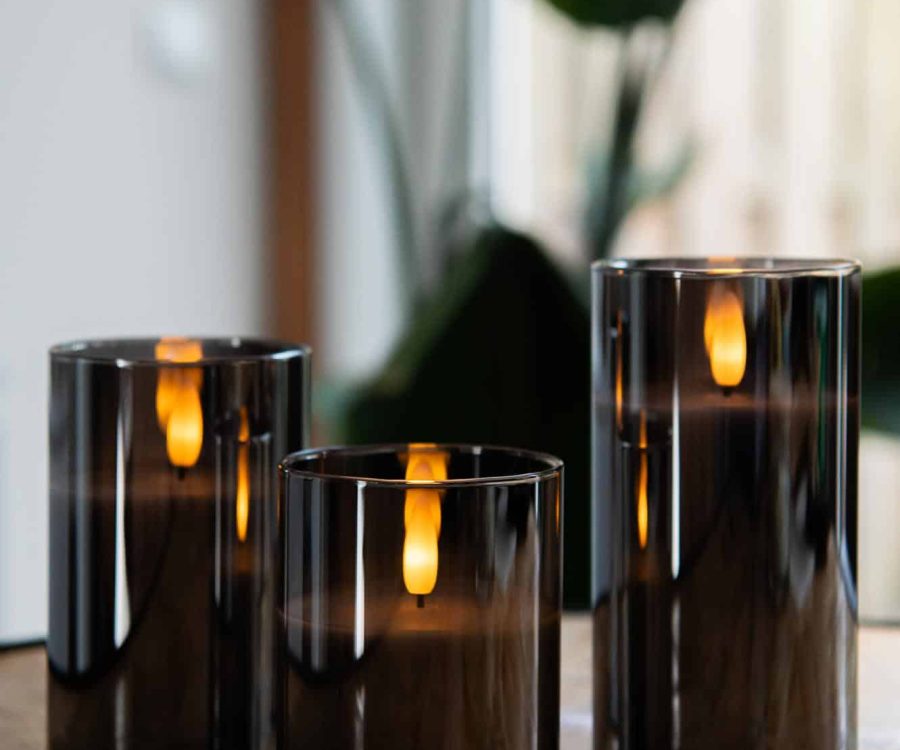 Three DORSETTO oplaadbare kaars in glas met 3D vlam staan op een houten oppervlak, hun sfeervolle licht weerkaatst in het glas. Op de achtergrond zorgen planten en daglicht voor de perfecte sfeer. Inclusief afstandsbediening, tot 105 uur brandtijd.