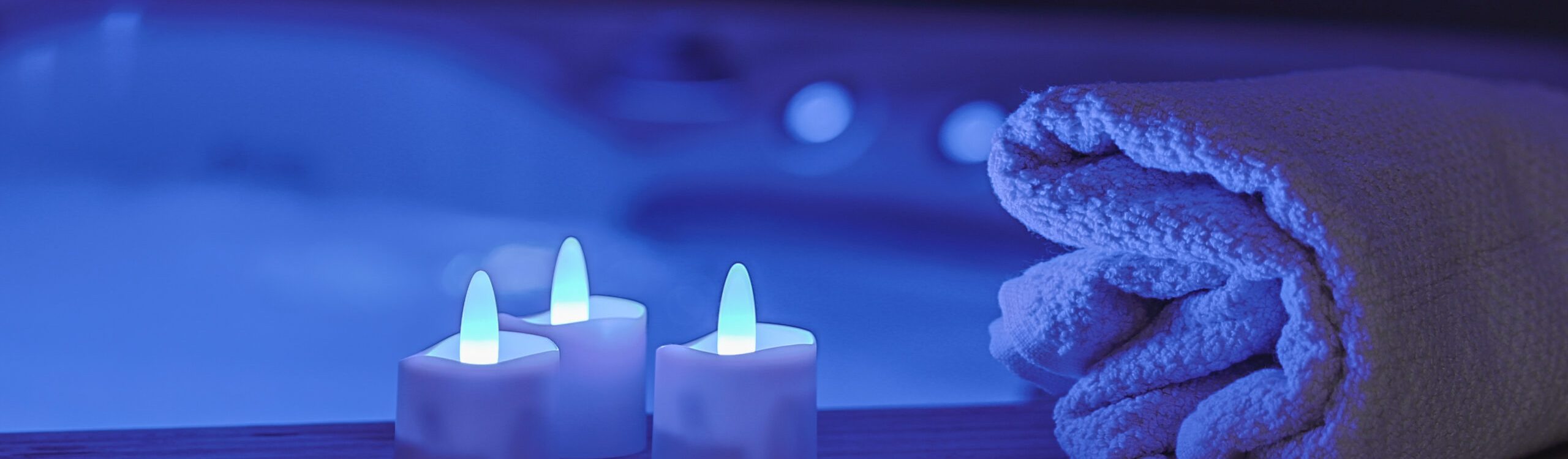 Een opgerolde handdoek en drie RGB LED-Waxine/theelichtjes oplaadbaar waterdicht (12 stuks) met afstandsbediening zitten op een houten plank boven een blauw verlicht bad en creëren een rustige, spa-achtige sfeer.