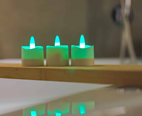 Drie kleine, witte RGB Led Waxinelichtjes oplaadbaar waterdicht (set van 12) met afstandsbediening gloeien groen op een houten dienblad, hun reflecties zichtbaar op het gladde oppervlak eronder tegen een zacht wazige achtergrond.