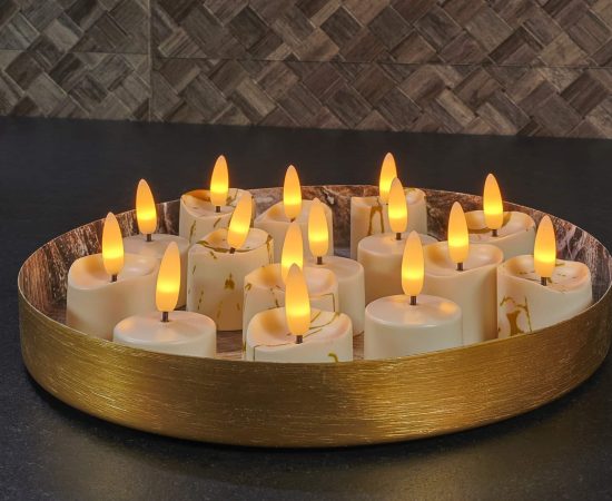 Een rond gouden dienblad bevat verschillende LED waxinelichtjes 3D brandtijd 175 uur oplaadbaar - met Afstandsbediening - 6 Stuks - timerfunctie, gerangschikt op een donker aanrecht met een tegelachterwand met textuur op de achtergrond.